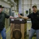Two men in branded shirts stand beside a large box labeled "GROW OPTIMIZED STEAM HUMIDIFIER" in a warehouse setting.