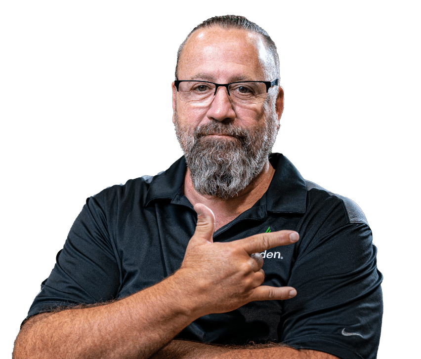 Un homme barbu, vêtu d'un polo noir, fait un geste de la main, porte des lunettes et affiche une expression confiante.