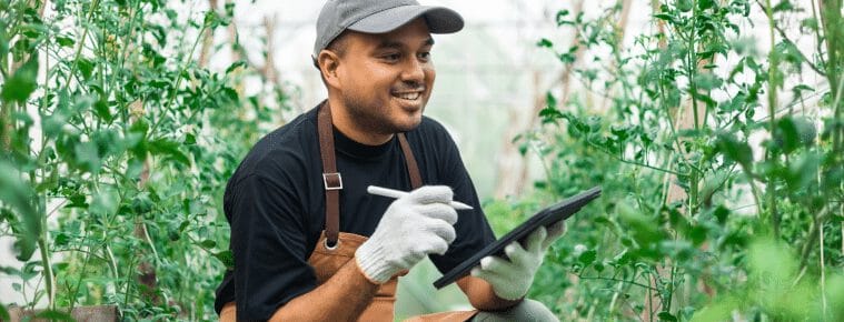 Jardinier souriant dans une serre, utilisant une tablette et un stylet, entouré de plantes vertes luxuriantes.