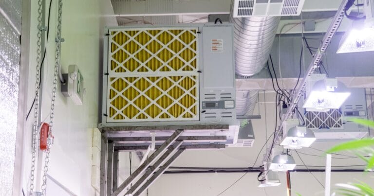 Suspended air conditioning unit with yellow filters, surrounded by ductwork and overhead lights in a modern facility.